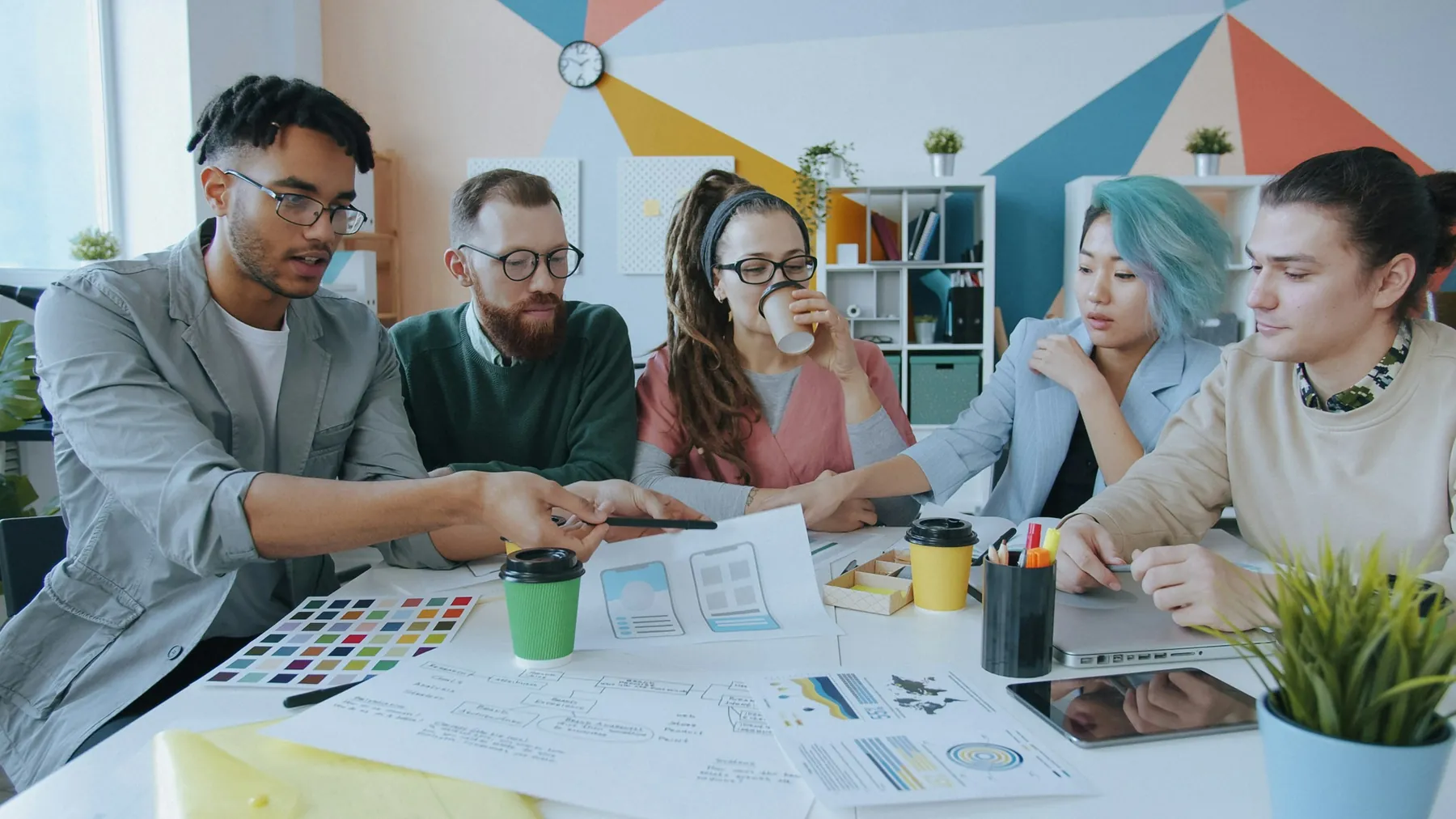 Five colleagues sit around a white table covered in printed layouts and colour swatches while one points at a page.